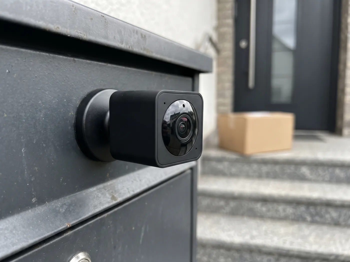 Kleine schwarze Mini-Kamera mit Magnetfuß an einer Metallfläche, im Hintergrund ein Paket vor der Tür